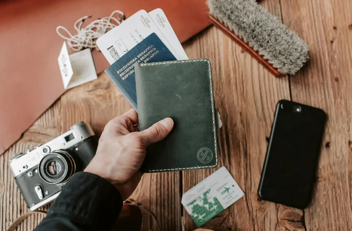 Homme tenant un carnet avec son passport et ses billets d'avion au dessus d'une table en bois ou l'on peut voir un appareil photo et un téléphone